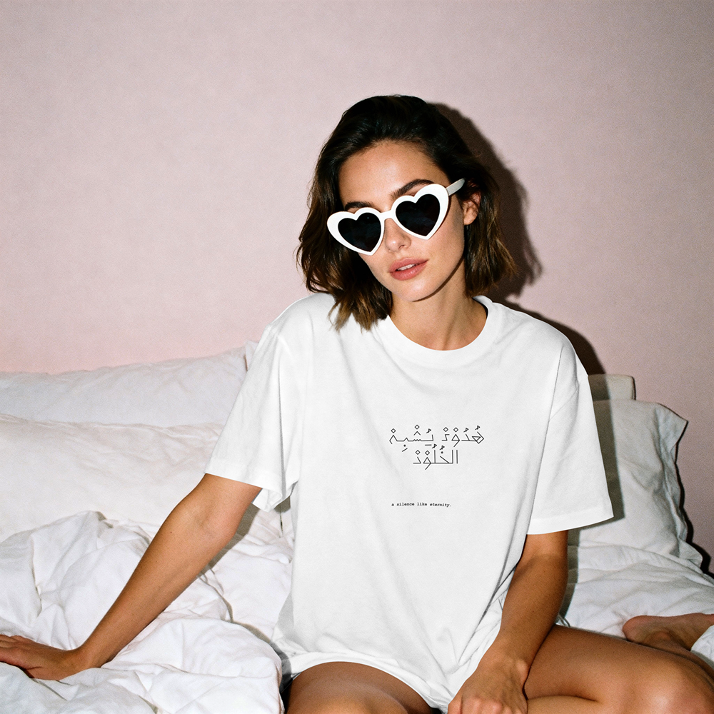 Woman wearing white graphic tee with Arabic text and heart sunglasses, sitting on bed