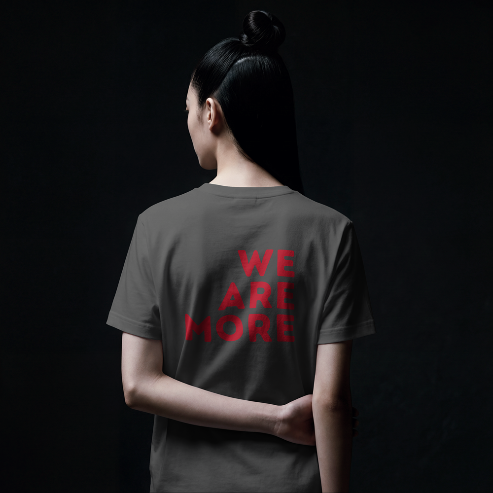 Woman wearing gray t-shirt with bold red 'WE ARE MORE' graphic print, back view, dark background